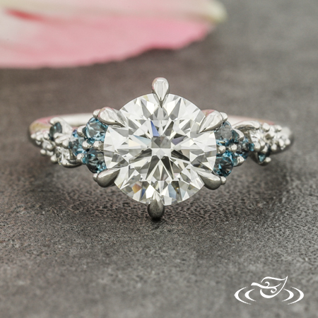 A round diamond in a six prong setting with accents of blue side stones set in a cluster arrangement.